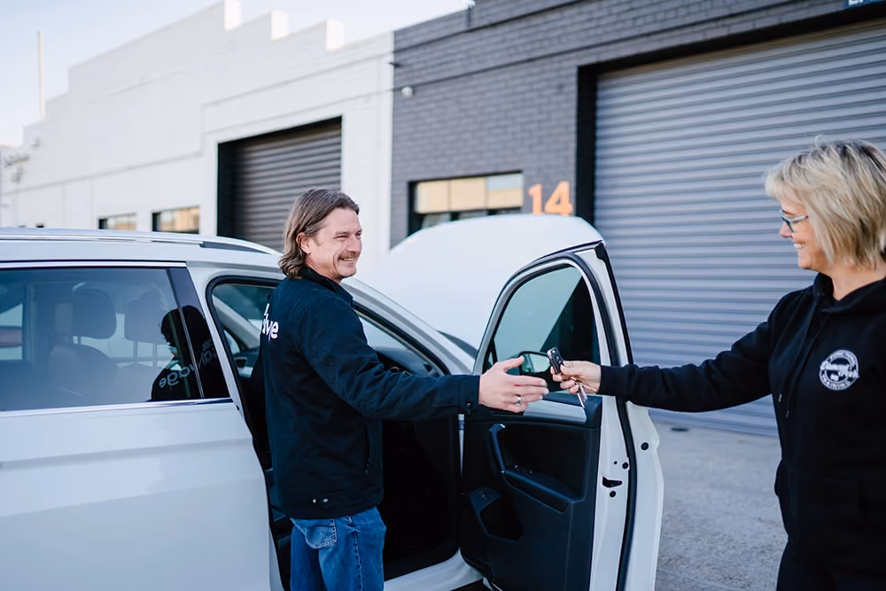Sell My Salvage employee receiving car keys from a customer beside a white vehicle outside an industrial workshop.