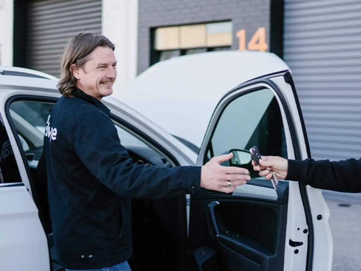A smiling man reaches out to take car keys from another person next to a white car with its bonnet open outside a workshop.