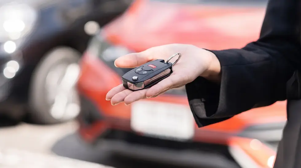 A person holding a black car key fob in their open palm, with a red car and a black car blurred in the background.
