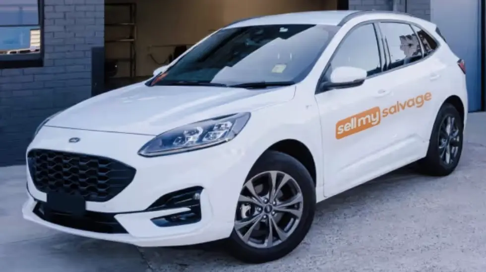 A white Ford SUV branded with orange 'sell my salvage' text parked outside a grey brick workshop with an open roller door.