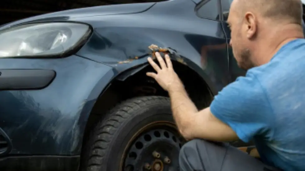 A man in a blue shirt crouching down to inspect dented, rusty damage on the front wing of a dark grey hatchback car.