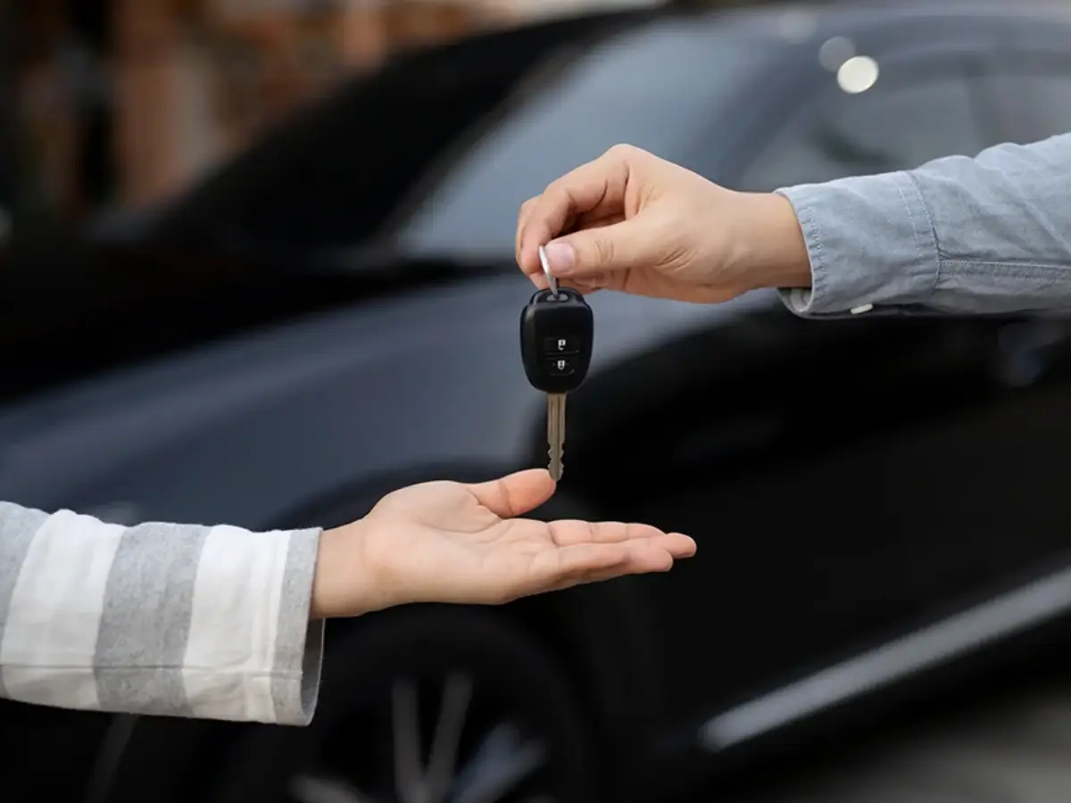 Close-up of a person handing over a black car key to another person's open palm in front of a sleek black vehicle.