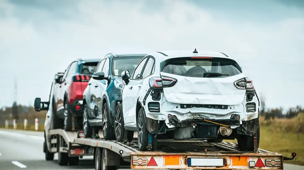 A car carrier trailer transporting several vehicles, with a white SUV featuring severe rear-end damage at the back.