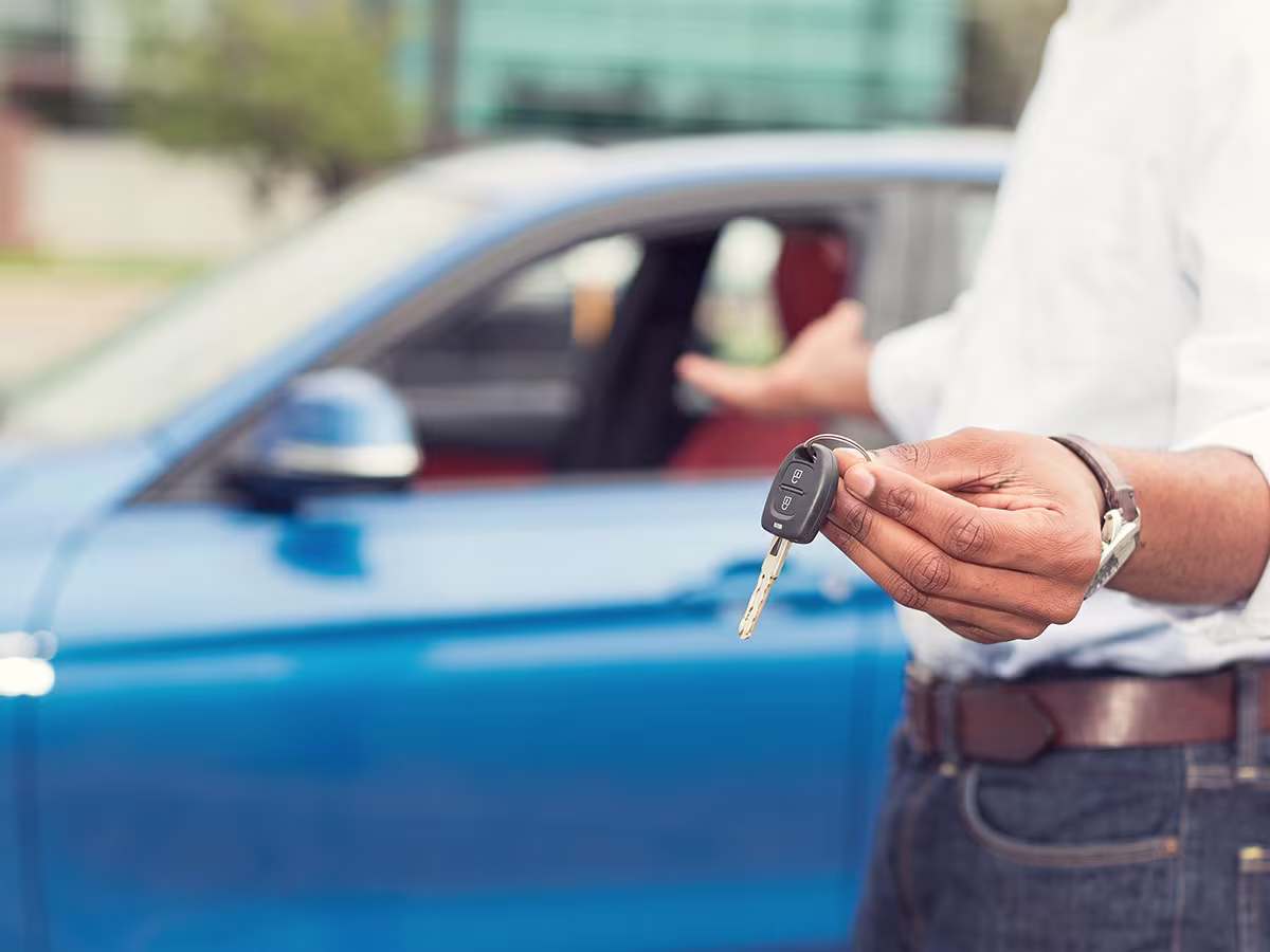 A person holding out a car key towards the camera while gesturing to a bright blue car parked behind them.