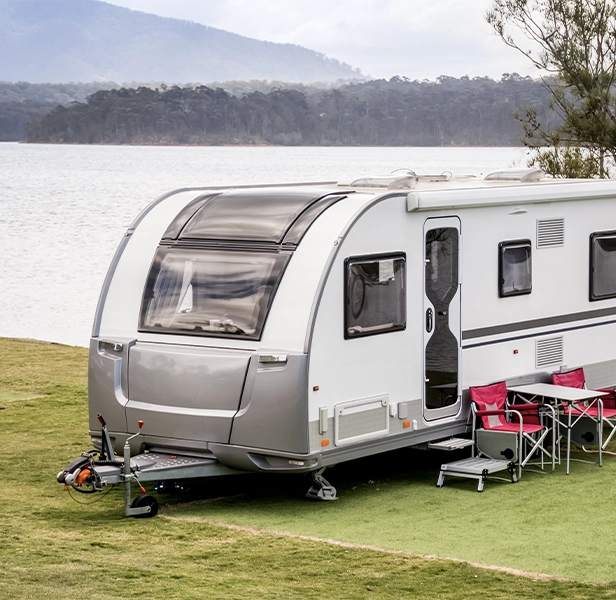 Caravan set up for camping near a lakeside with chairs and table outside.