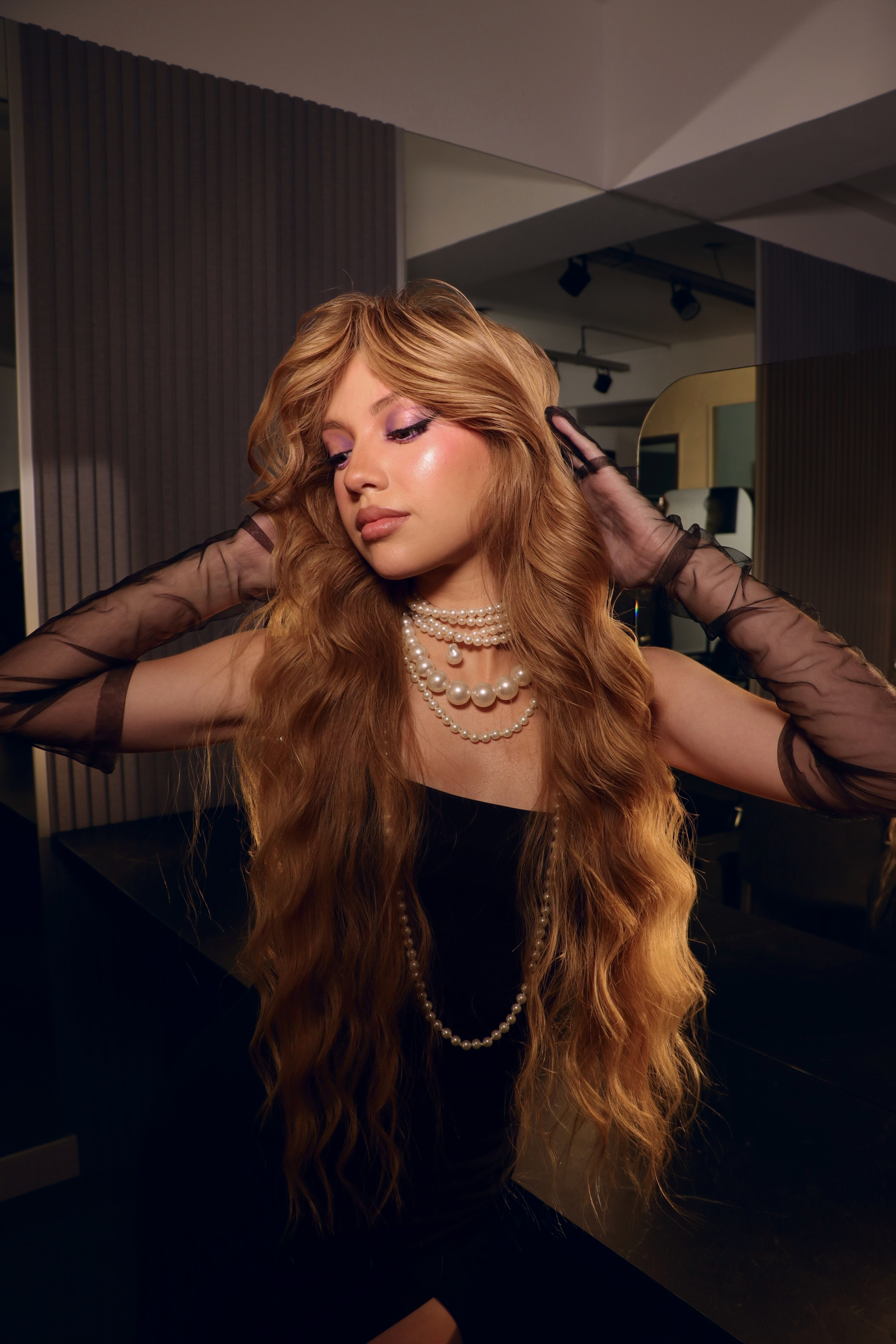 Woman with long wavy blonde hair wearing layered pearl necklaces, sheer black gloves, and a black dress, posing with eyes closed.