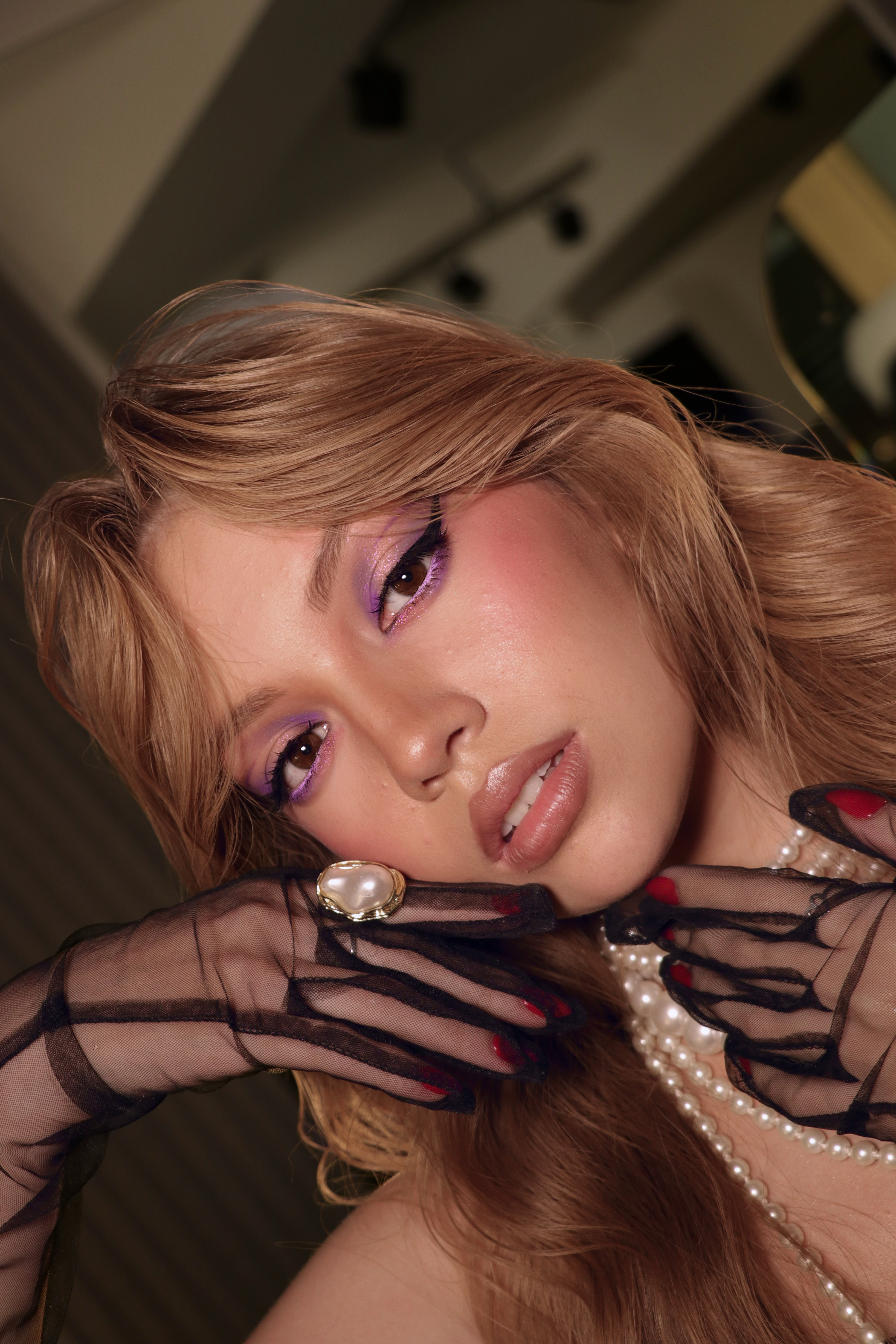 Close-up of a woman with purple eyeshadow and winged eyeliner wearing sheer black gloves with red nails, a large pearl ring, and a multi-strand pearl necklace.