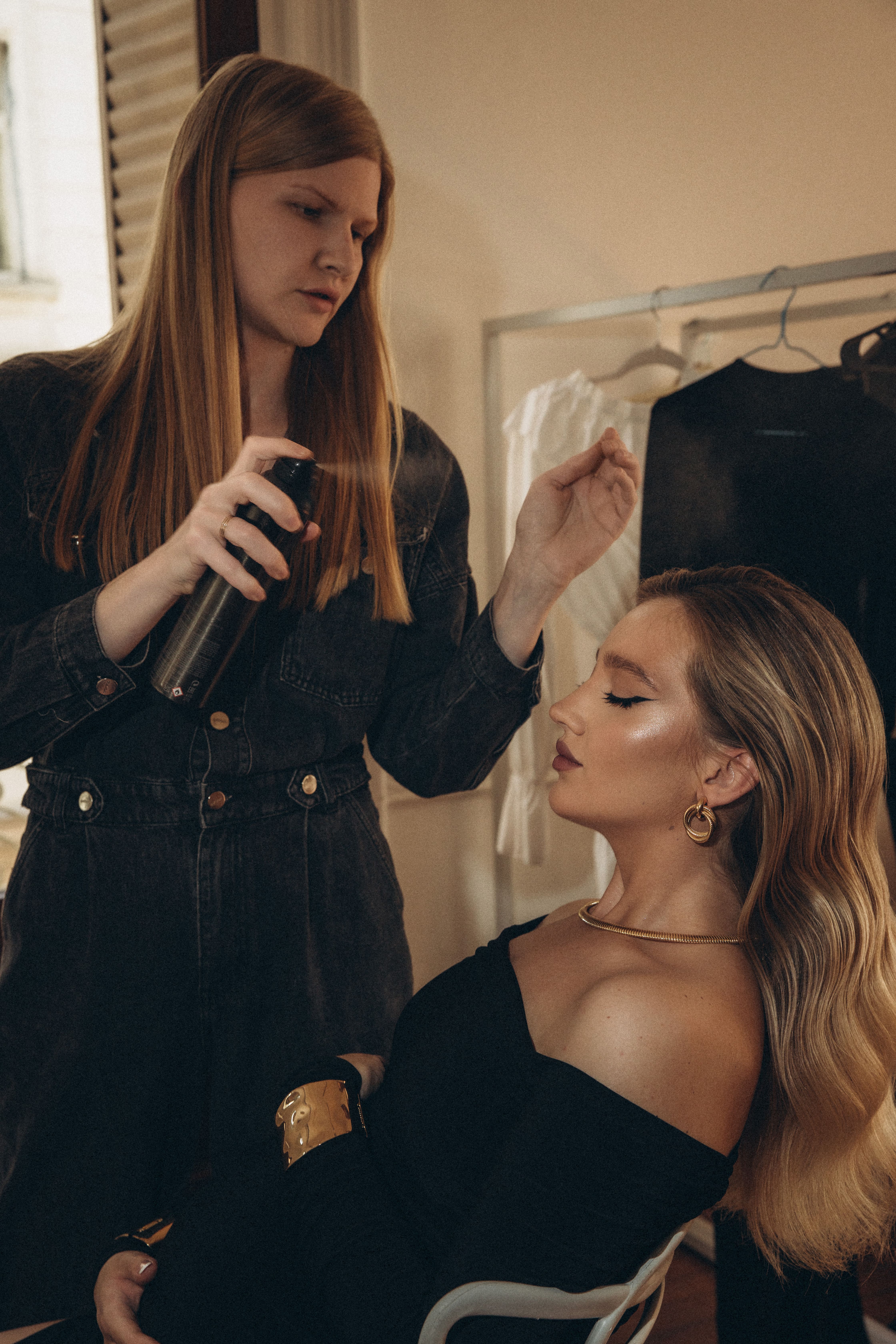 A hairstylist sprays hair product onto a seated woman with styled blonde waves and elegant makeup.