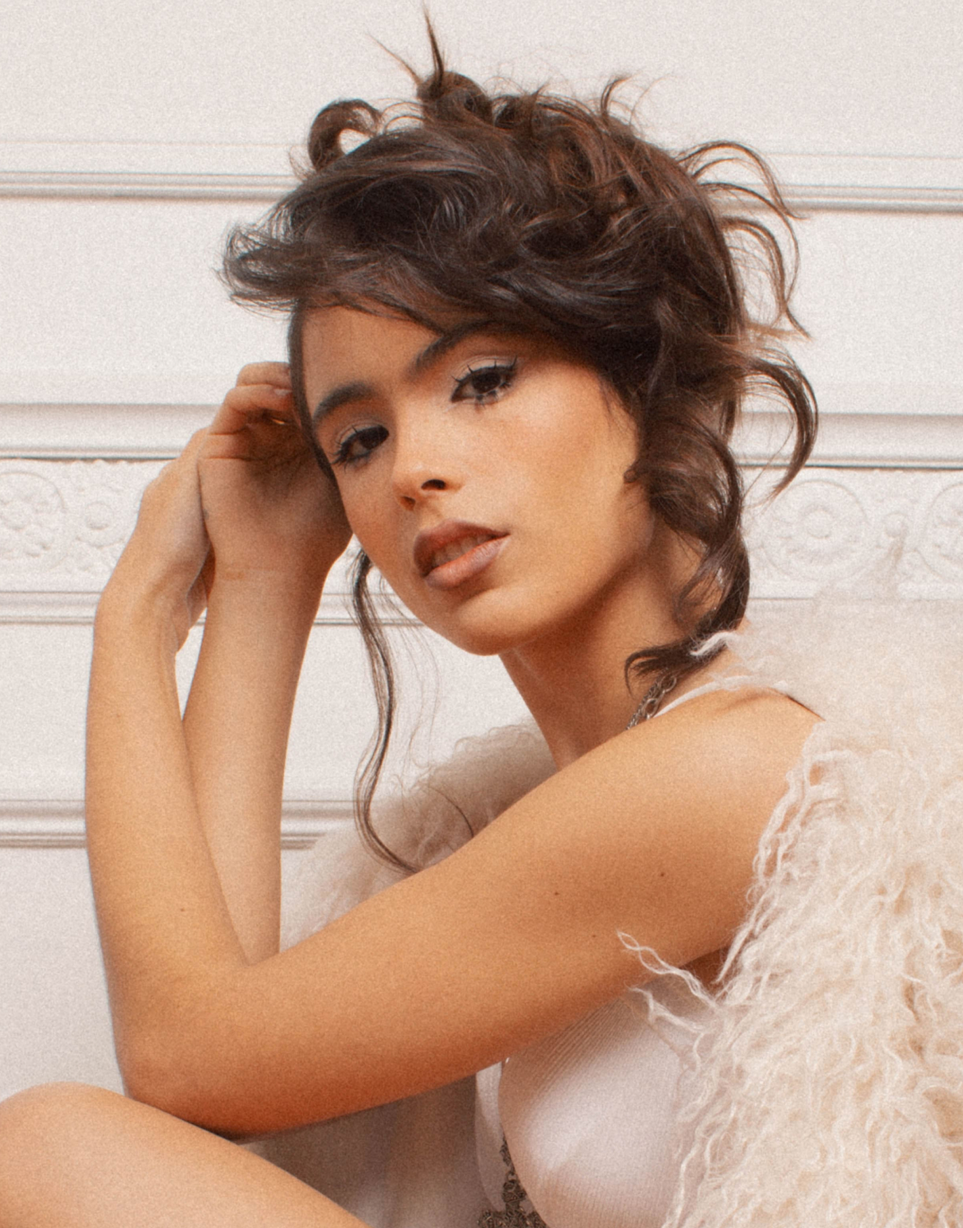 Mujer joven con vestido blanco y abrigo peludo claro en cuclillas sobre piso de madera en interior elegante.