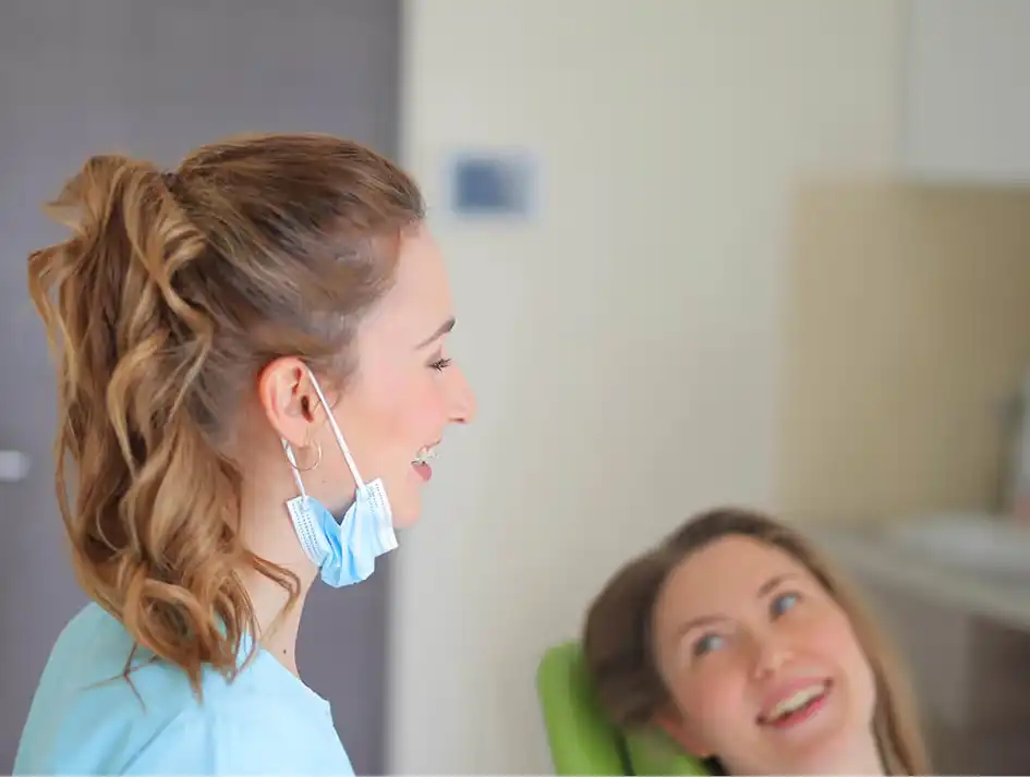 Eine lächelnde Zahnärztin in Maske und eine lachende Patientin, die in einen Spiegel blickt.
