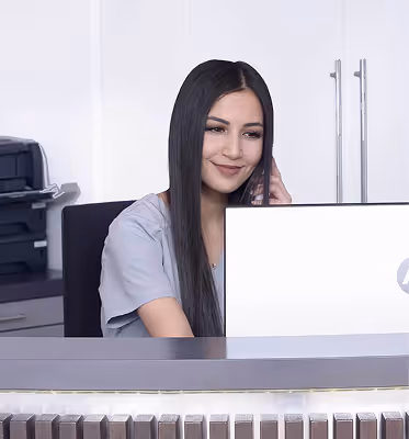 Smiling woman with long dark hair sitting at a desk using a laptop.