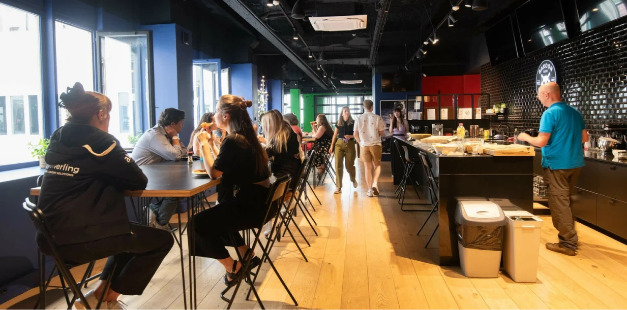People sitting and eating at a long table in a bright, modern communal dining area with wooden floors and large windows.