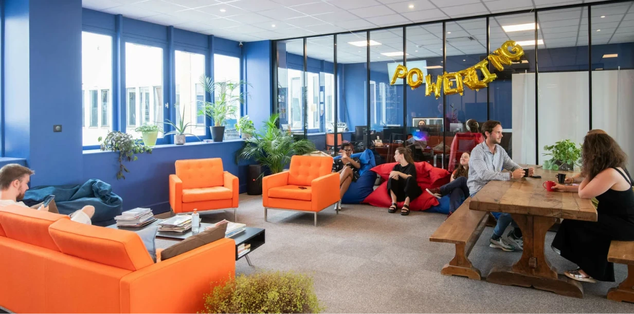 Office lounge with orange sofas and armchairs, people sitting on bean bags and wooden benches, and gold balloon letters spelling POWERING.