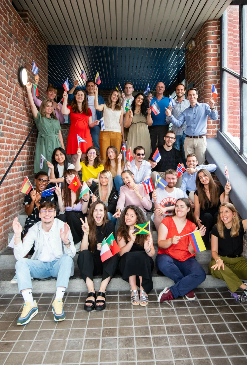 Group of diverse young adults smiling and sitting on stairs, holding various international flags in a brick-walled indoor space.
