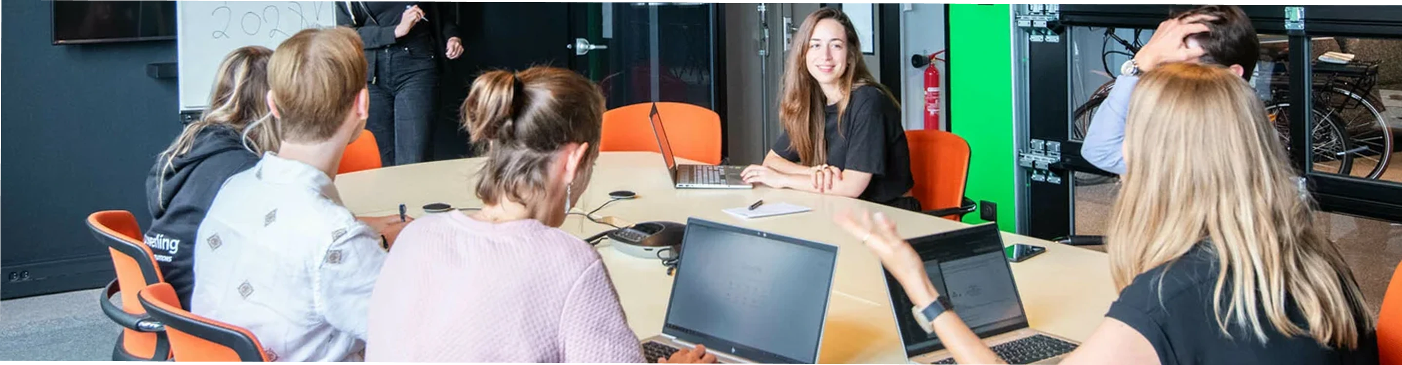 Group of six young adults sitting around a conference table in an office, some using laptops, engaged in discussion.