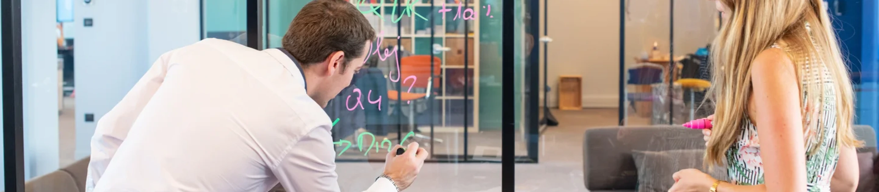 Man and woman collaborating by writing colorful notes on a glass board in a modern office.