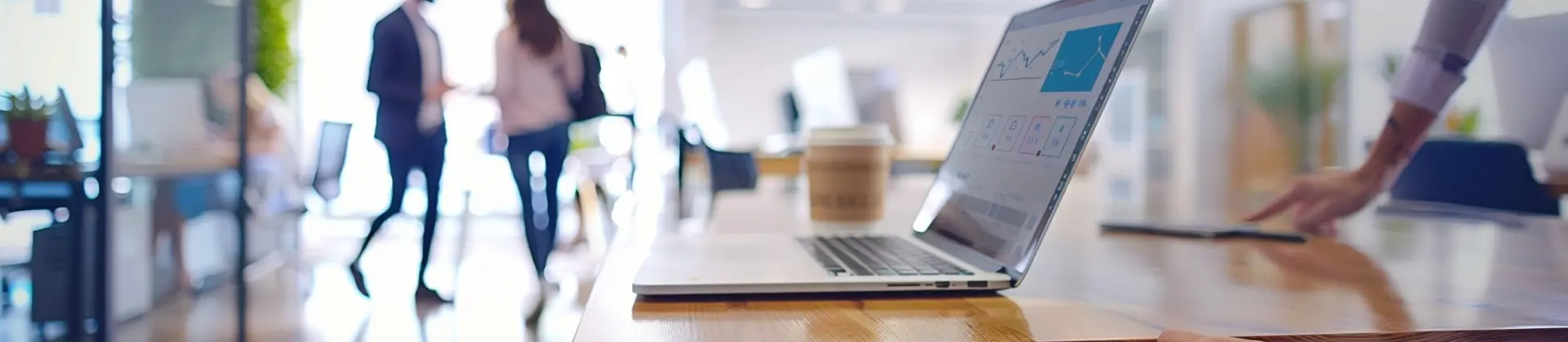 Laptop on a wooden table displaying data charts, with office workers blurred in the background.