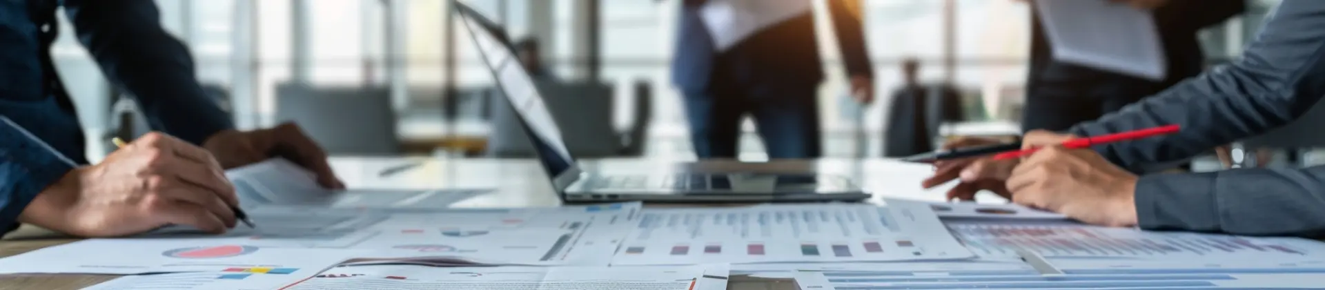 Two people reviewing business charts and documents on a table with a laptop in a bright office.