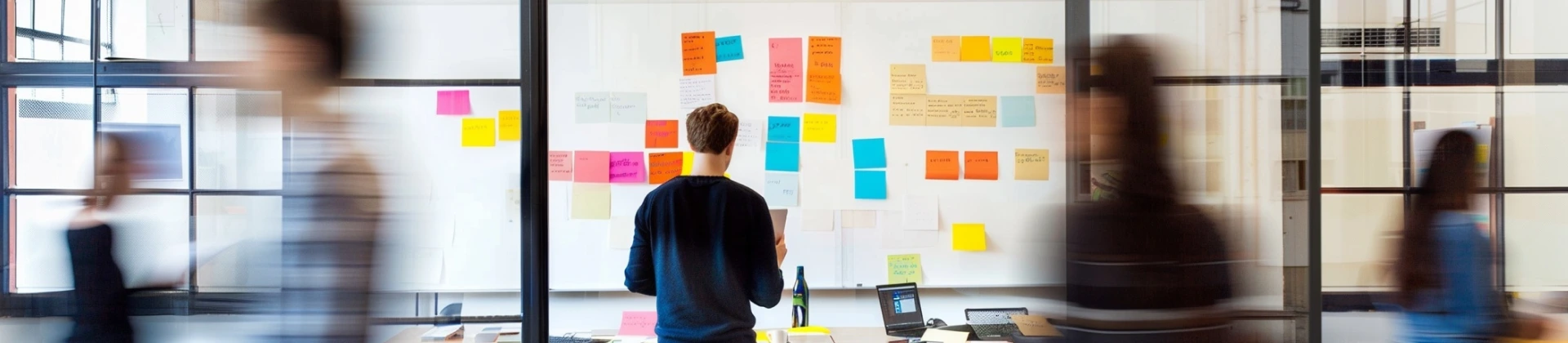 Person standing in front of a whiteboard covered with colorful sticky notes in a modern office, with blurred figures walking past glass walls.