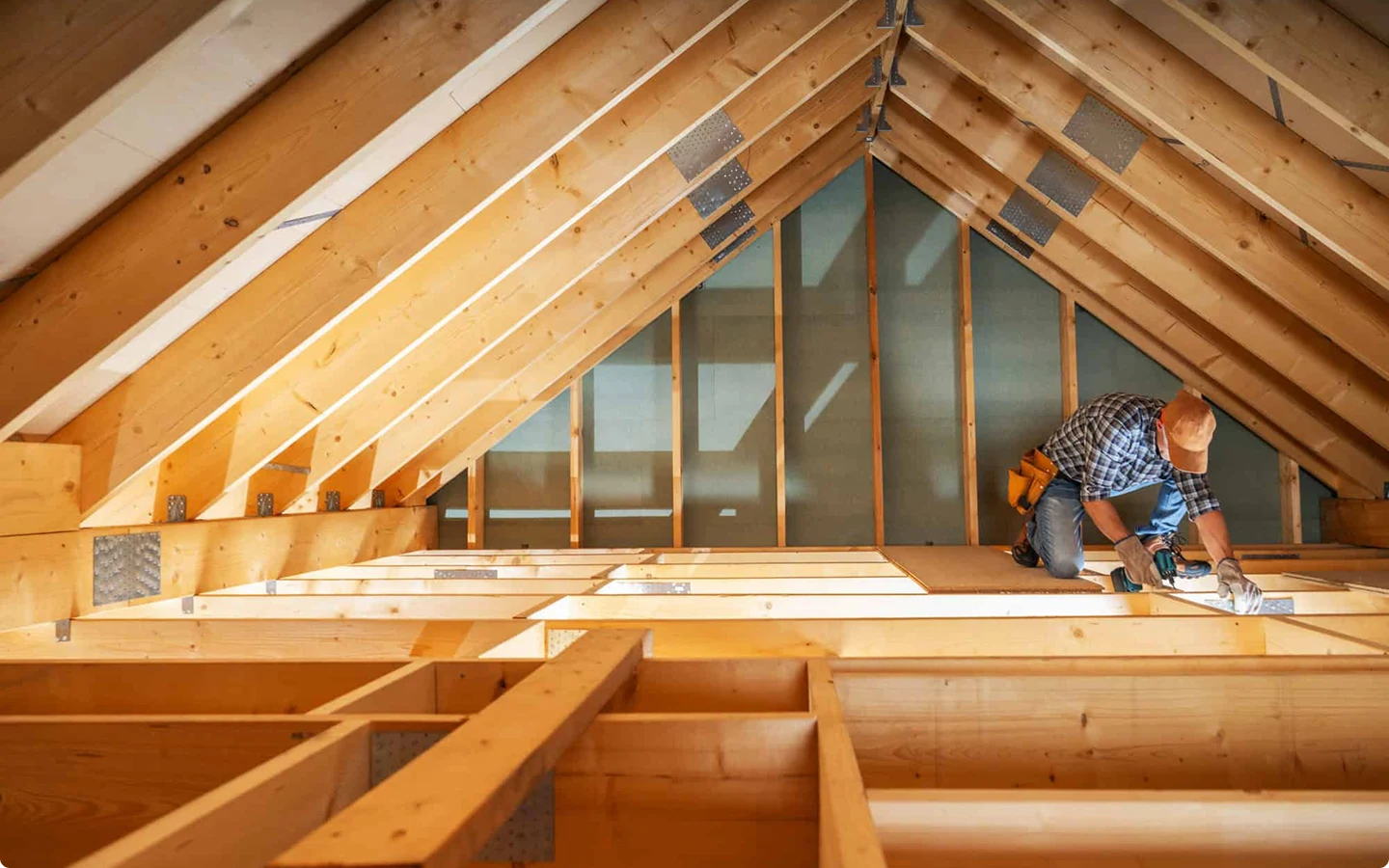 Worker in a cap drills wooden beams inside an attic under construction with exposed rafters.