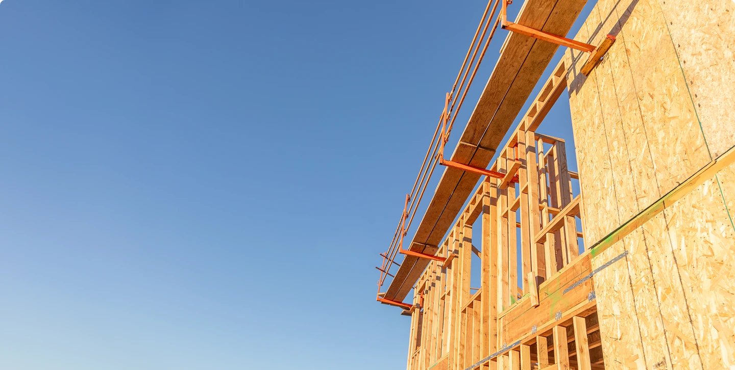 Wooden frame of a building under construction with scaffolding against a clear blue sky.