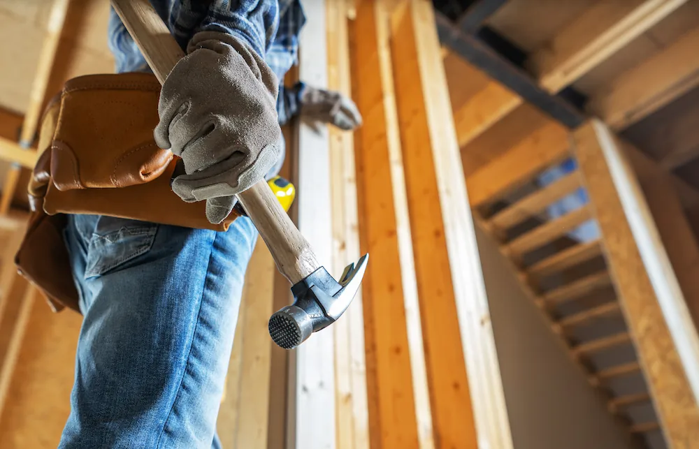 Construction worker wearing gloves and tool belt holding a hammer inside a wooden structure.