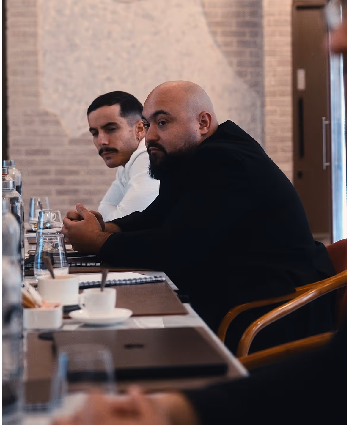 Deux hommes assis à une table de réunion avec des carnets et des tasses devant eux.
