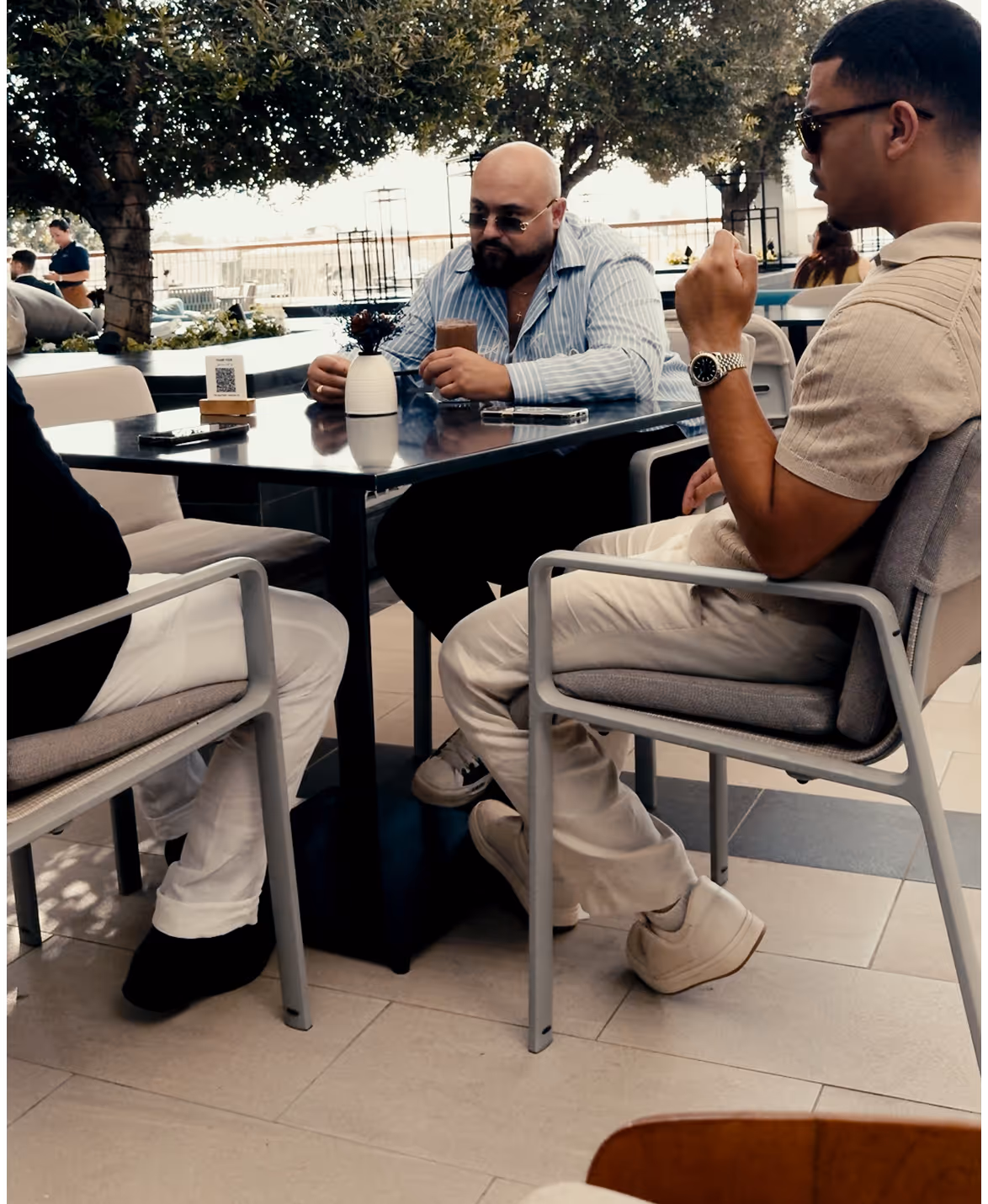 Trois hommes assis à une table de café en plein air sous des arbres, discutant et tenant des boissons.