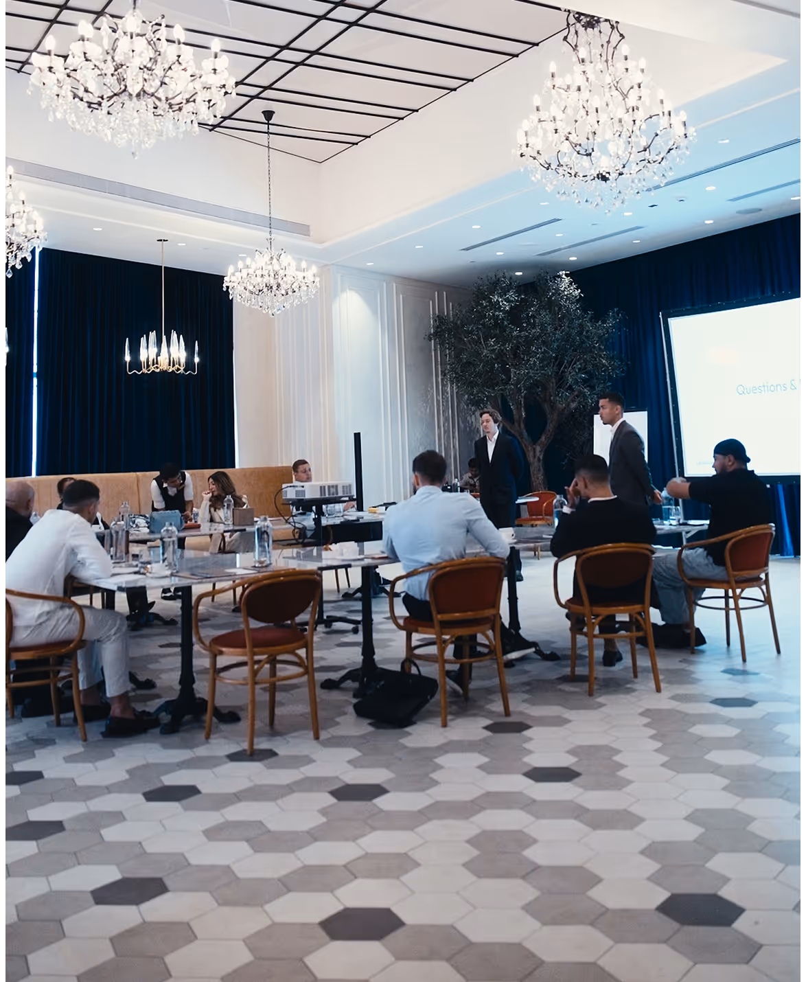 Salle de réunion élégante avec sol en carreaux hexagonaux, plusieurs personnes assises autour d'une table et une présentation projetée à l'avant.