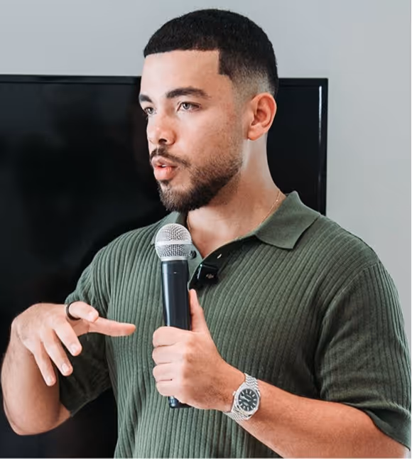 Homme avec polo vert tenant un microphone et parlant devant un écran noir.