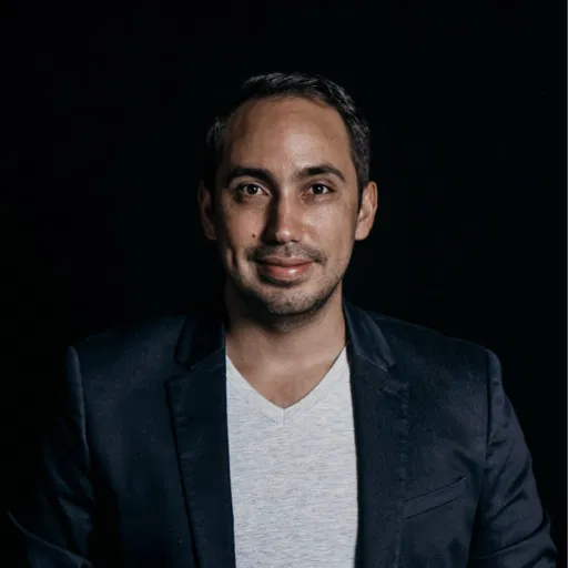 Portrait d'un homme souriant portant une veste noire et un t-shirt blanc sur fond sombre.