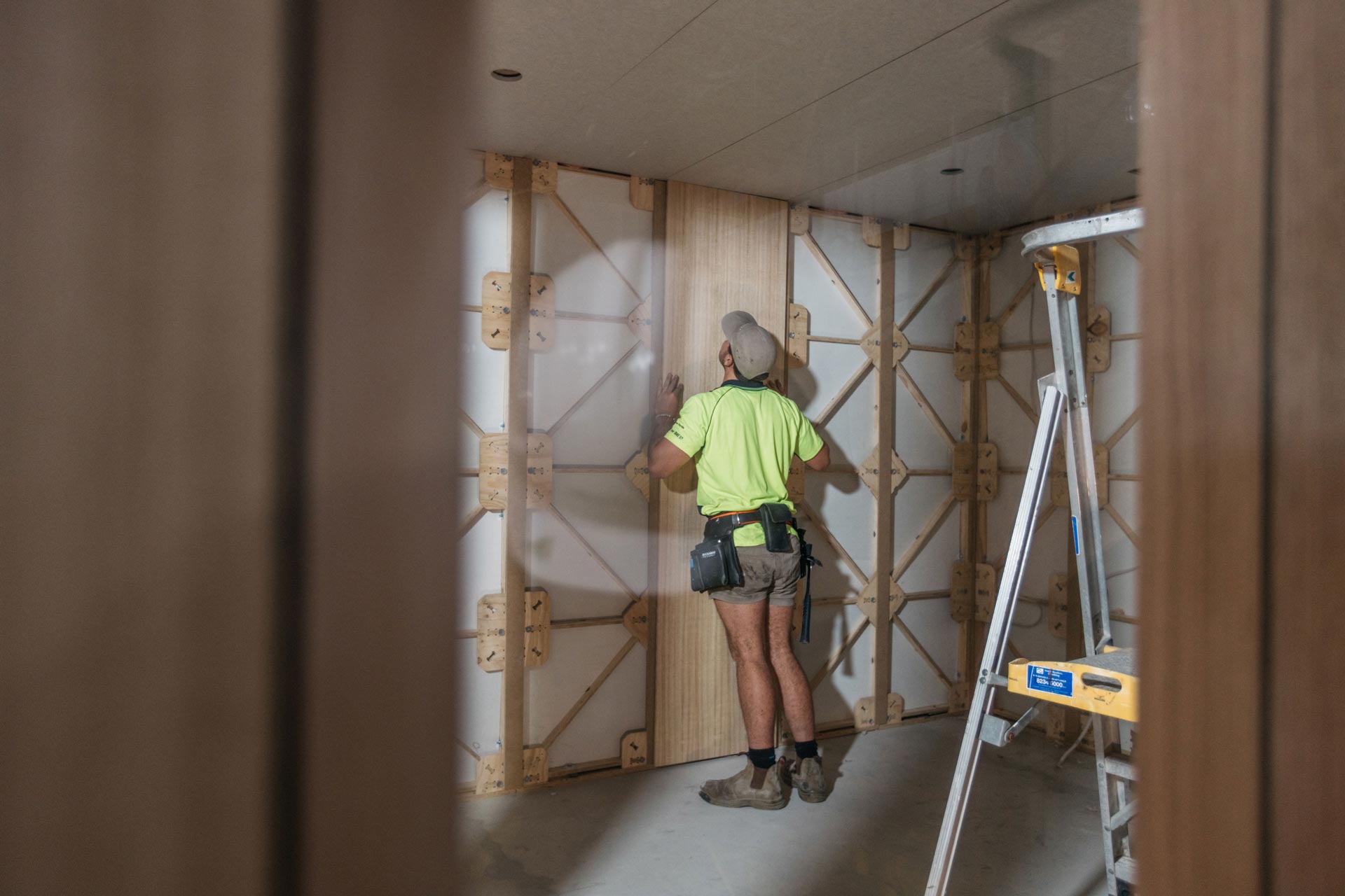 A builder wearing a high vis t-shirt, tool belt and some steel cap boots, putting on a Tasmanian Oak Lining on an XFrame Panel in an XFrame room.