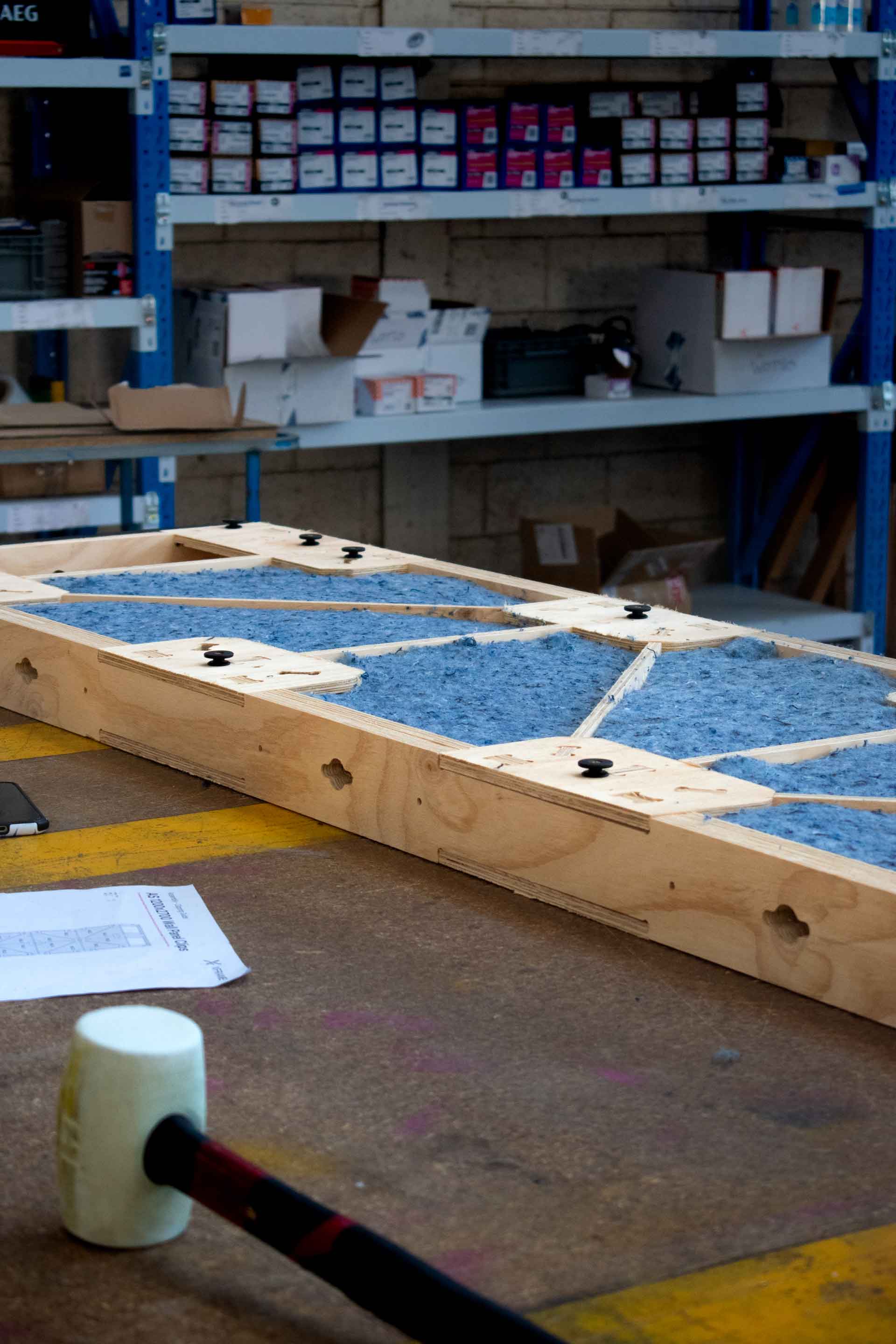 A 600mm XFrame panel with denim insulation on a work bench in a factory with a hammer in the foreground and shelved in the background.