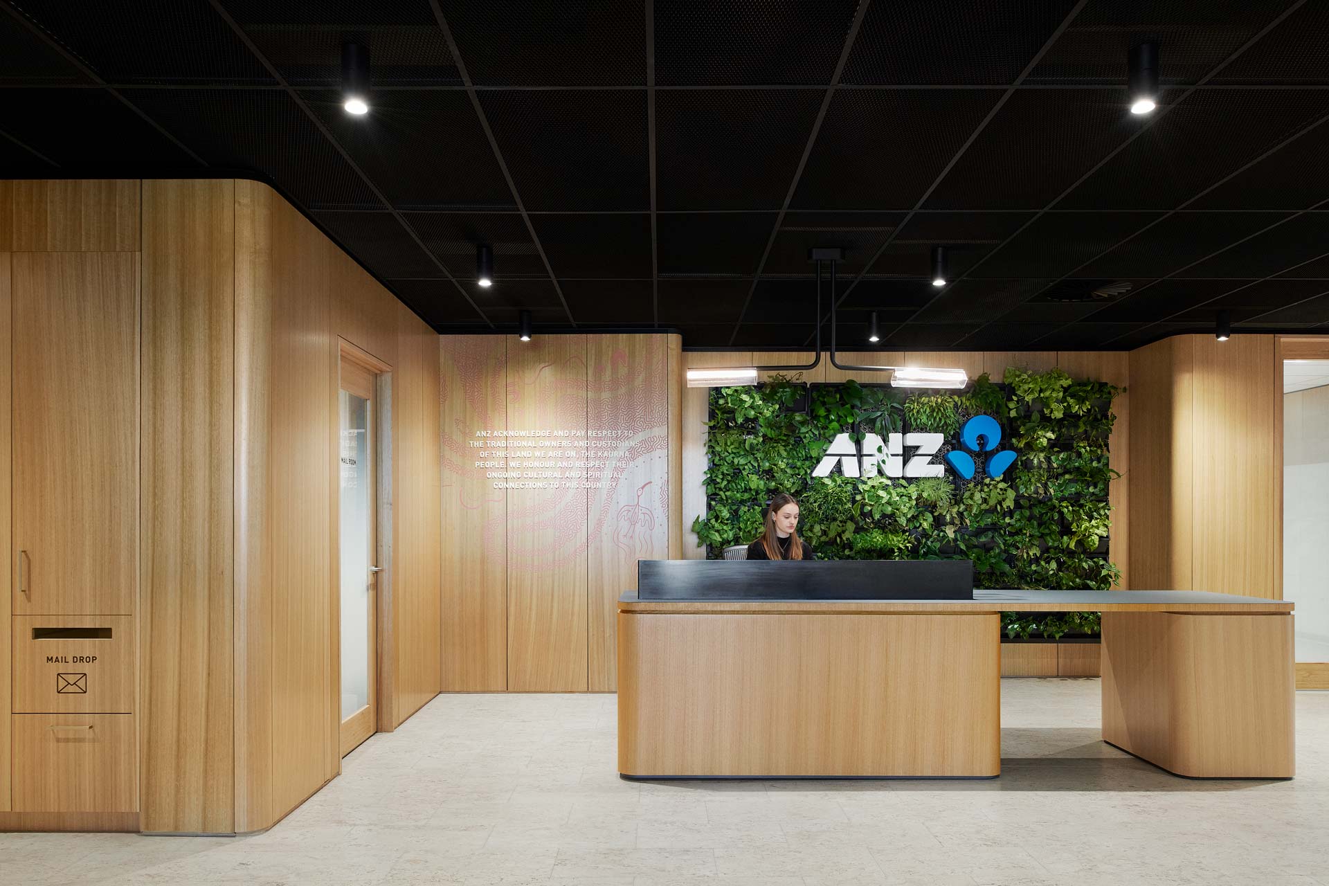 ANZ Piri St office reception desk, with a lady behind it, with a ANZ logo on a green plant feature wall. and meeting rooms two the left and right.