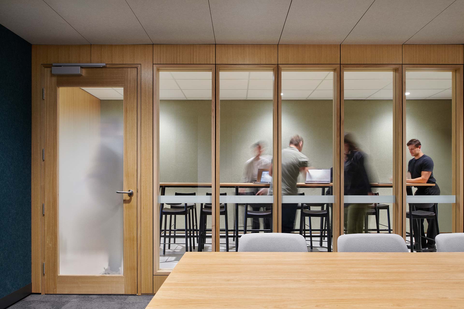 Looking into a meeting room through the window front, with a tall table and tall chairs with people on their laptops.