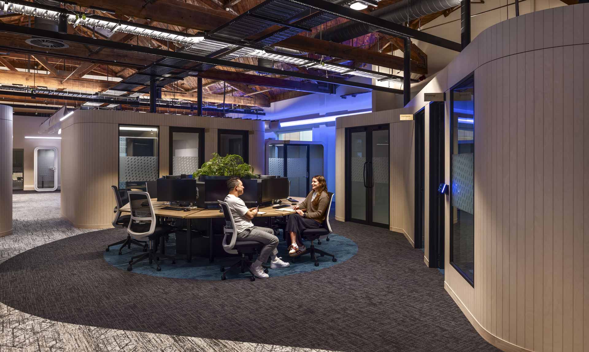 Meridian Energy office space with a circle of desks with computers and chairs, with a man and women sitting on office chairs, chatting together. They are surrounded by meeting rooms with curved corners, and birch wooden slat linings.