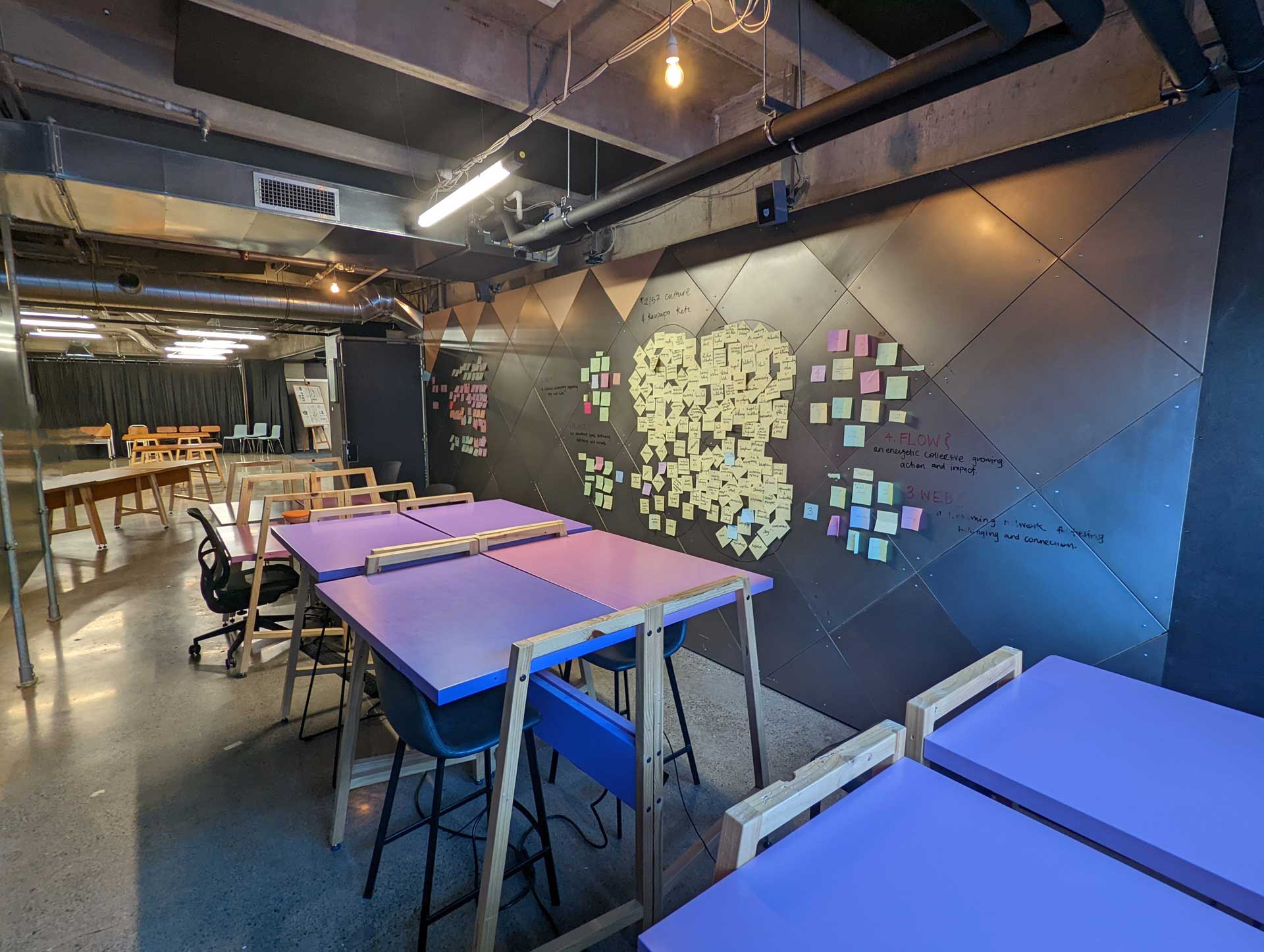 A wall made out of XFrame panels with steel diamond linings, with post in notes and whiteboard markers on it, with purple and pink, high and low desks with chairs.