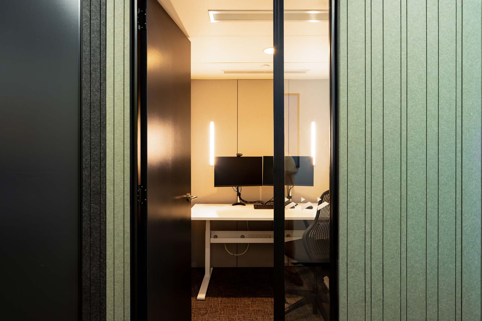Looking into a Booster meeting room through an opened black door, with a white desk, two monitors, a office chair with PET interior linings and PET grooved green exterior linings.