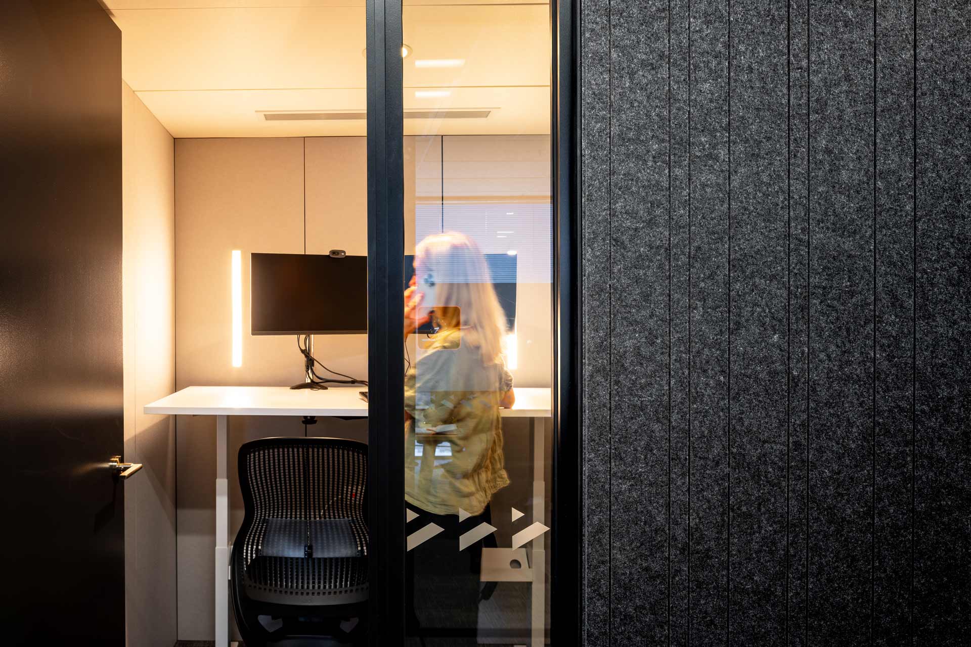 A girl in a green t-shirt on the phone, inside a Booster meeting room, with a standing desk, two monitors and a desk chair, with PET interior linings, and PET dark grey grooved exterior linings and a black door.