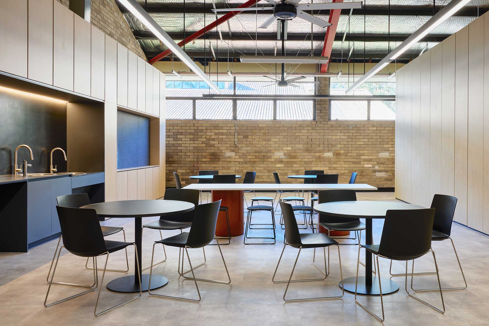 Breakout space with four black round tables and black chairs, with a long timber tables with orange pillar legs in the middle. With a mini kitchenette on the left and a side of a room on the right.
