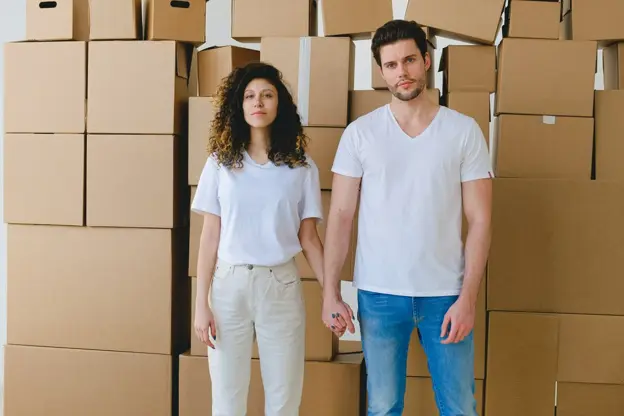 Young couple holding hands in front of stacked cardboard moving boxes, preparing to move into or out of their home.
