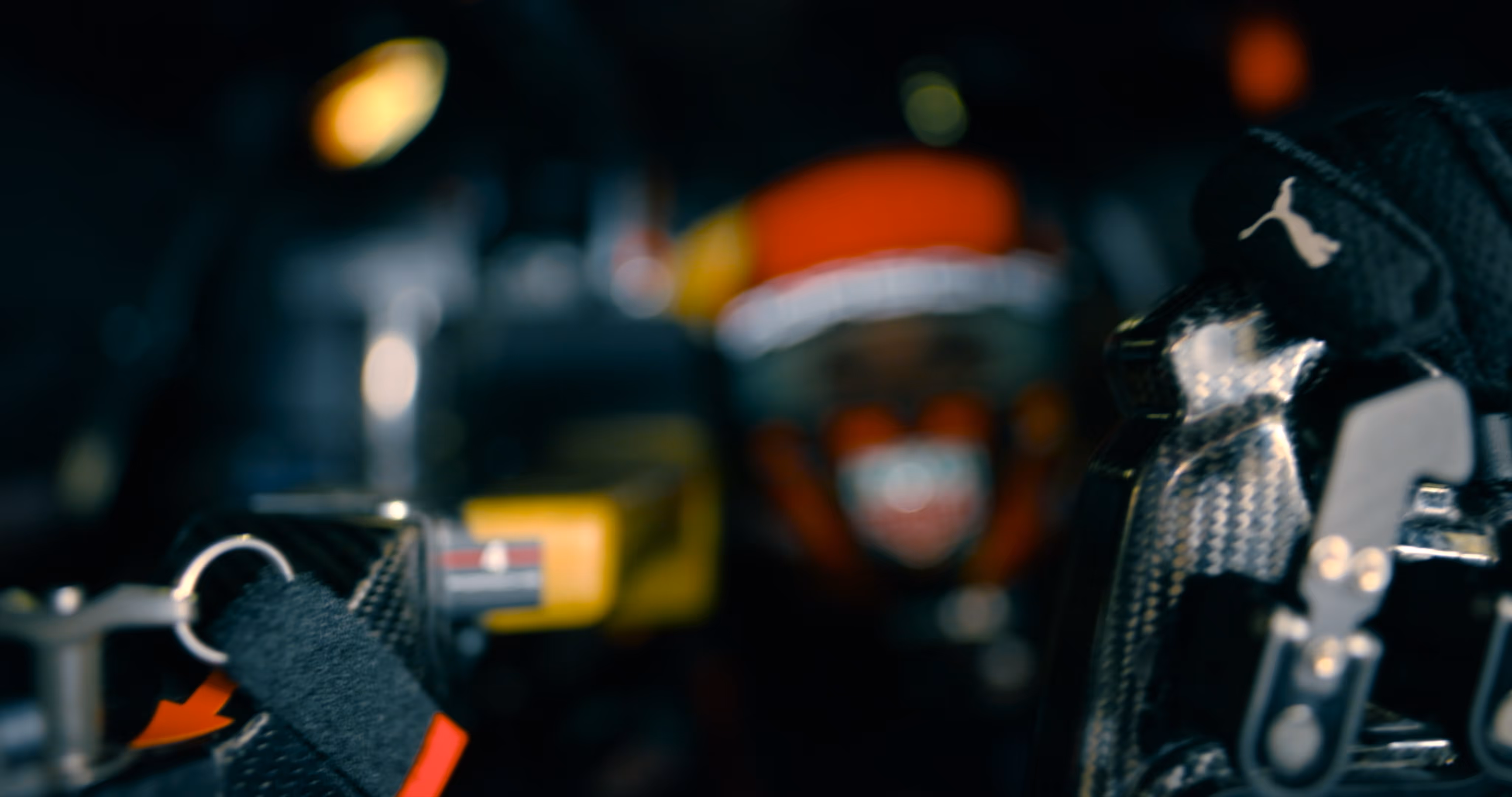 Close-up of a carbon fiber racing seatbelt harness with a blurred race car driver wearing a helmet in the background.