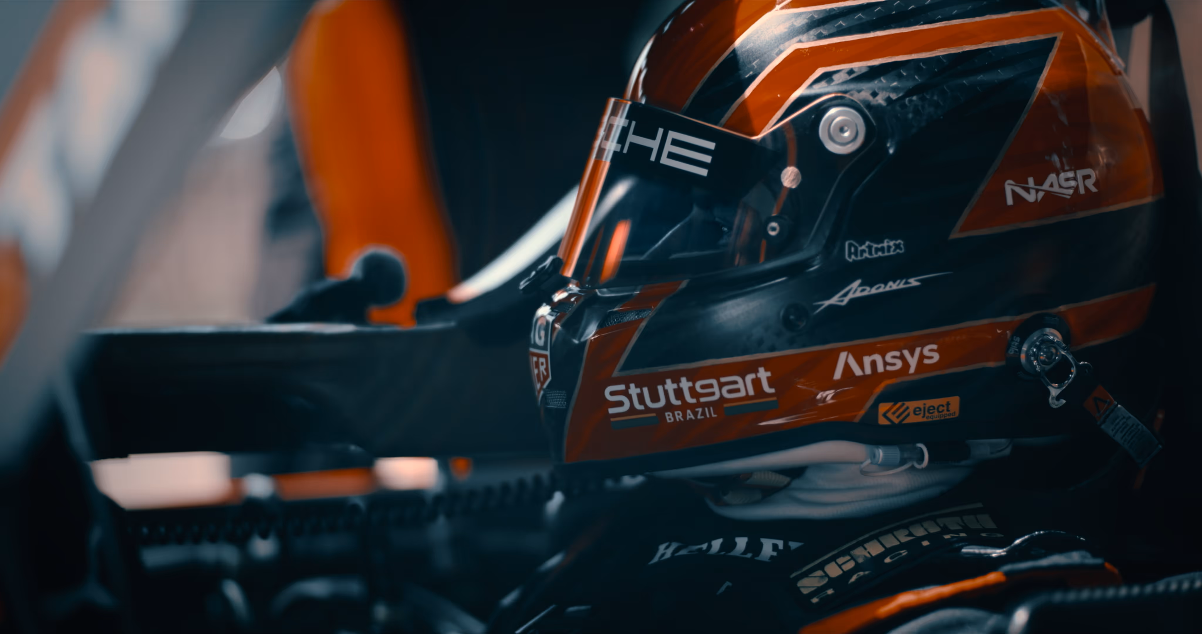 Close-up of a race car driver wearing a black and orange helmet with sponsorship logos inside a race car cockpit.