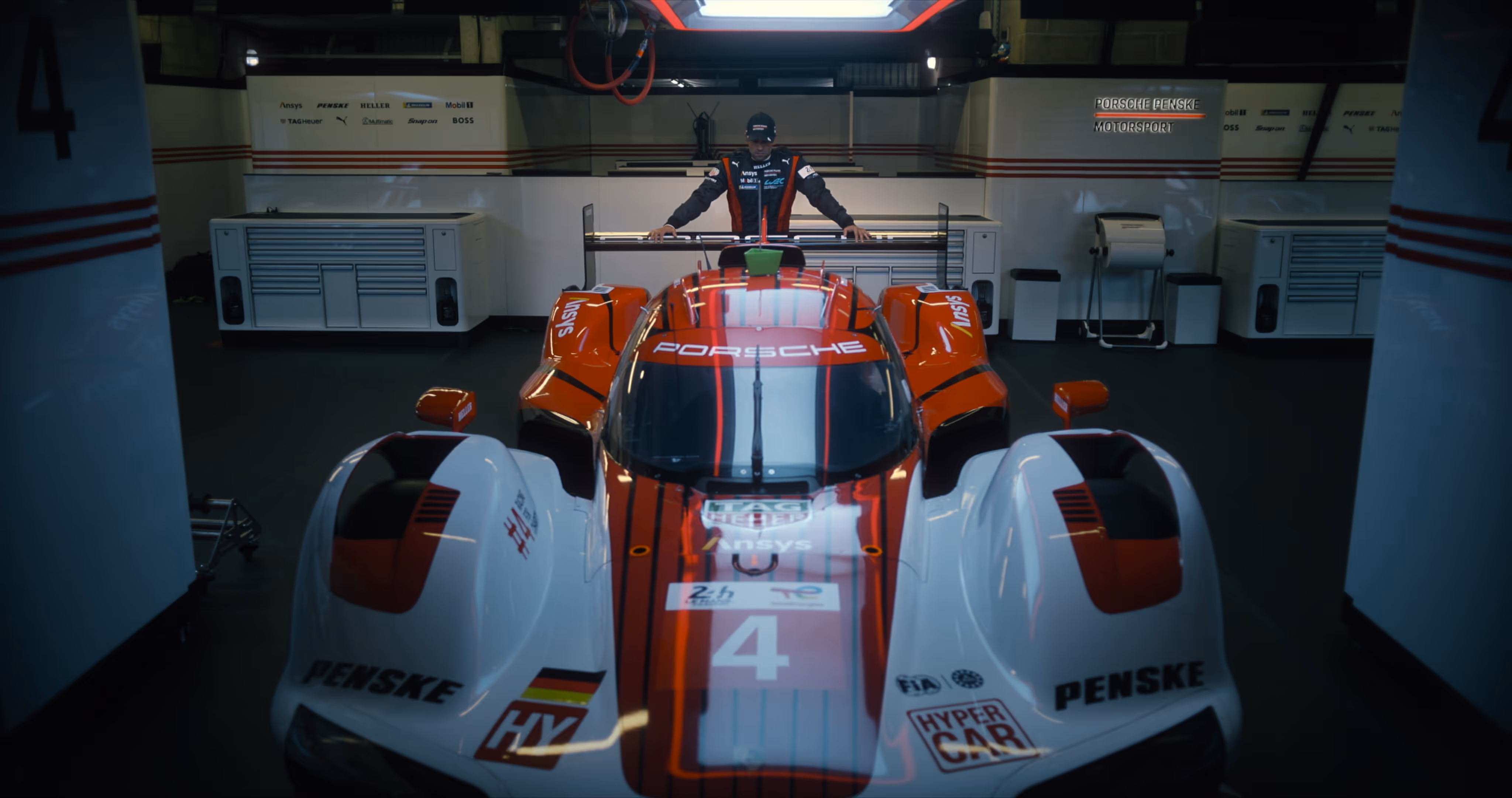 Driver standing behind a white and red Porsche Penske Hypercar number 4 in a garage.