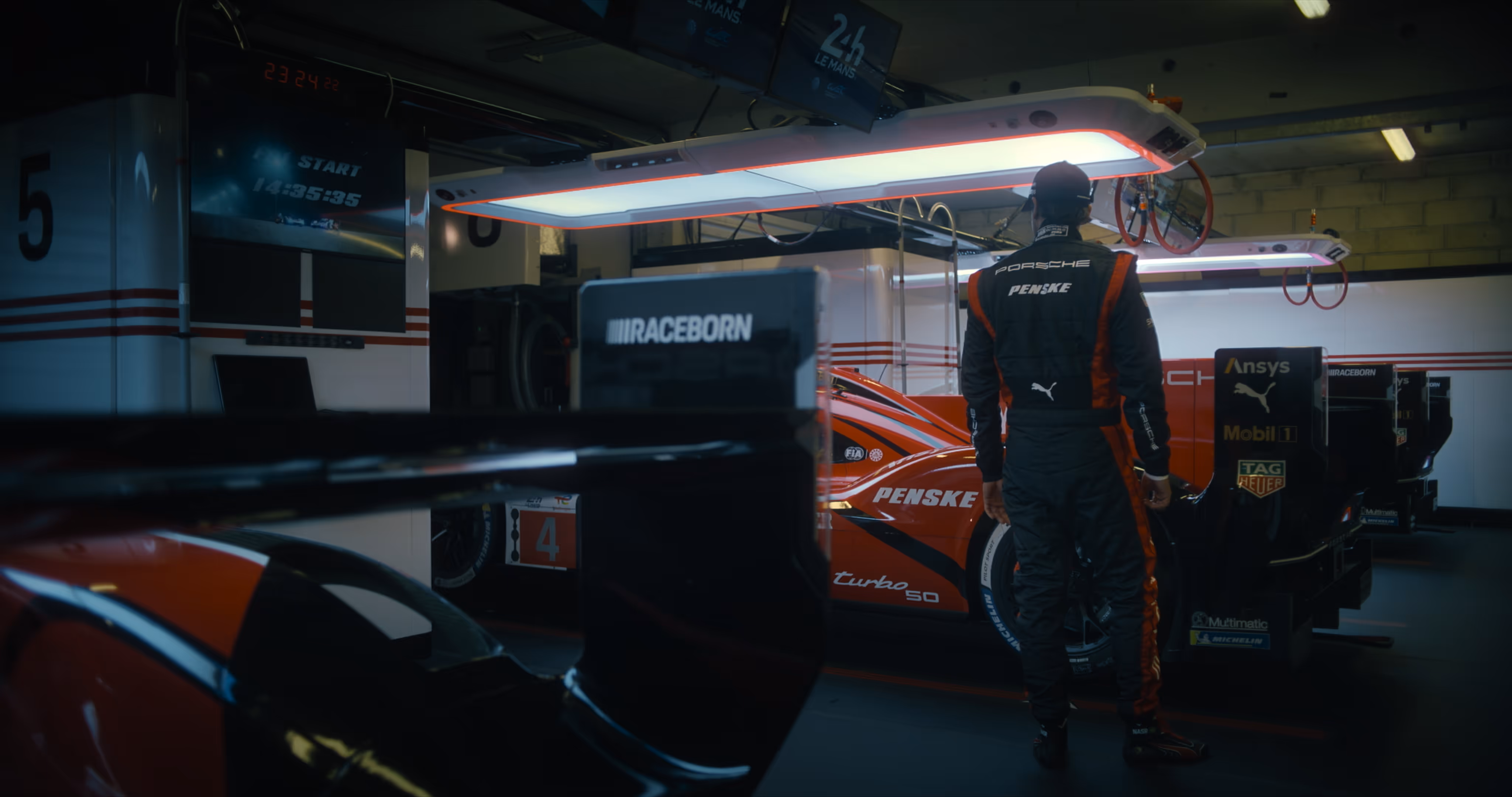 Race car driver standing next to a Porsche Penske race car in a garage pit area under bright overhead lights.