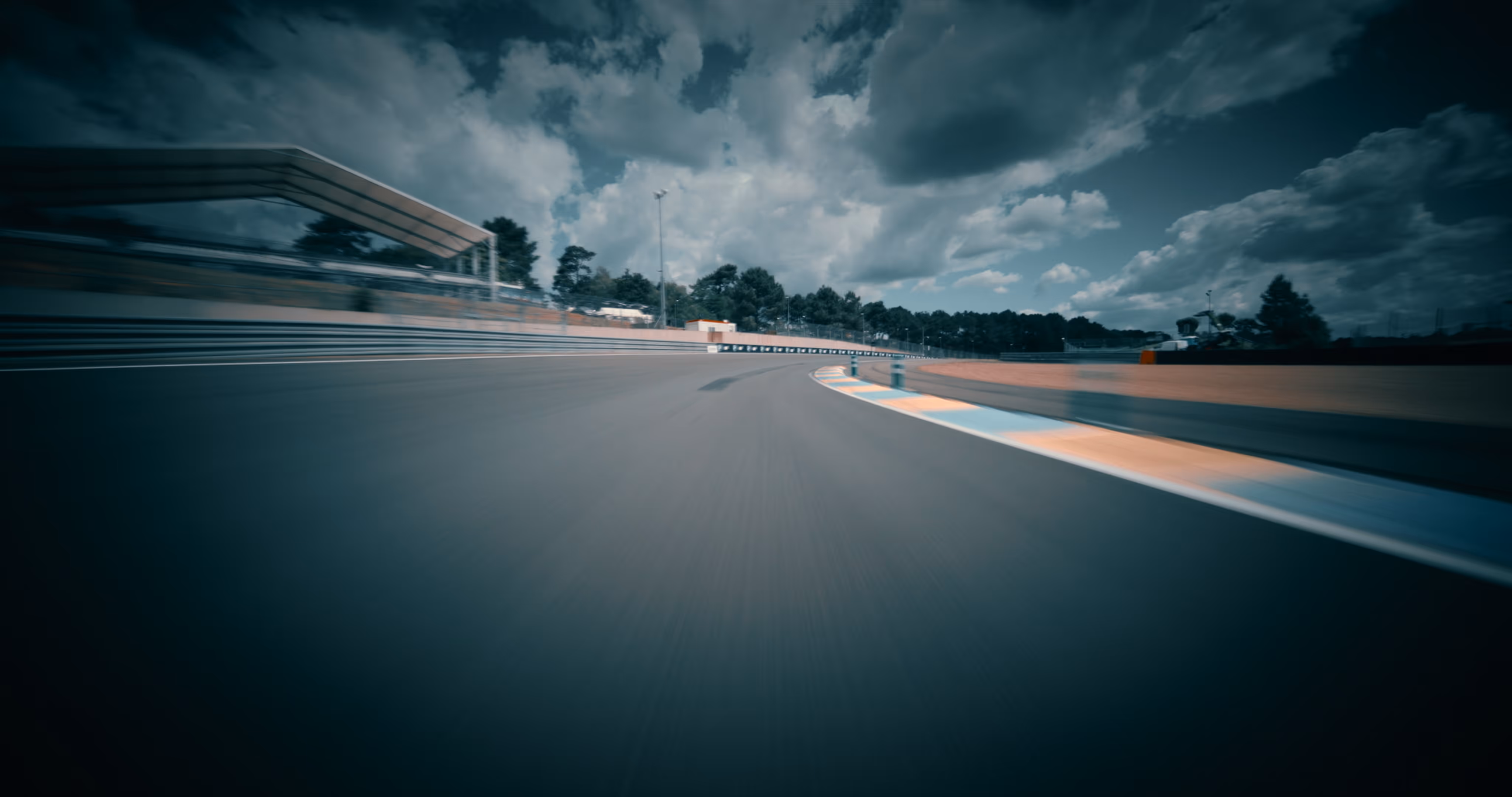 Empty racetrack with a sharp curve under a cloudy sky surrounded by trees and barriers.