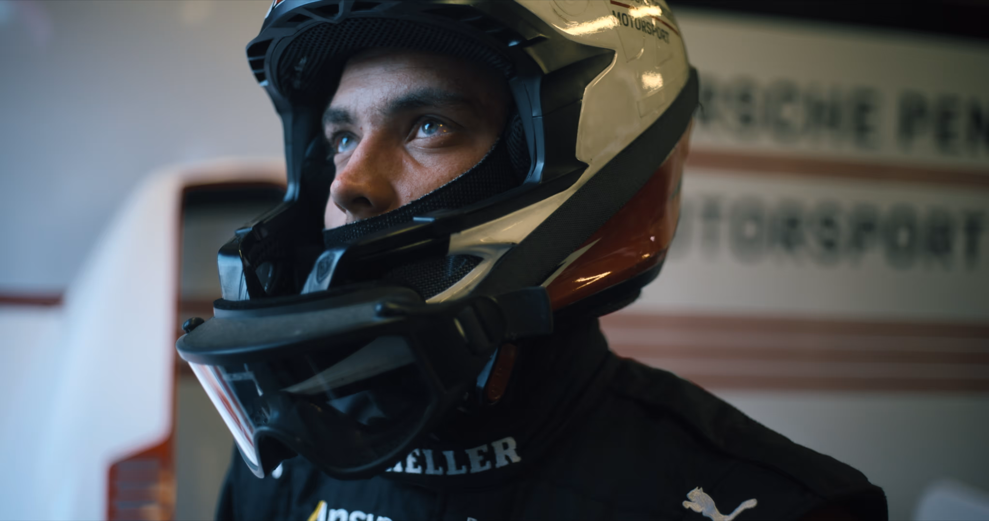 Close-up of a race car driver wearing a white and red helmet and black racing suit looking upward.