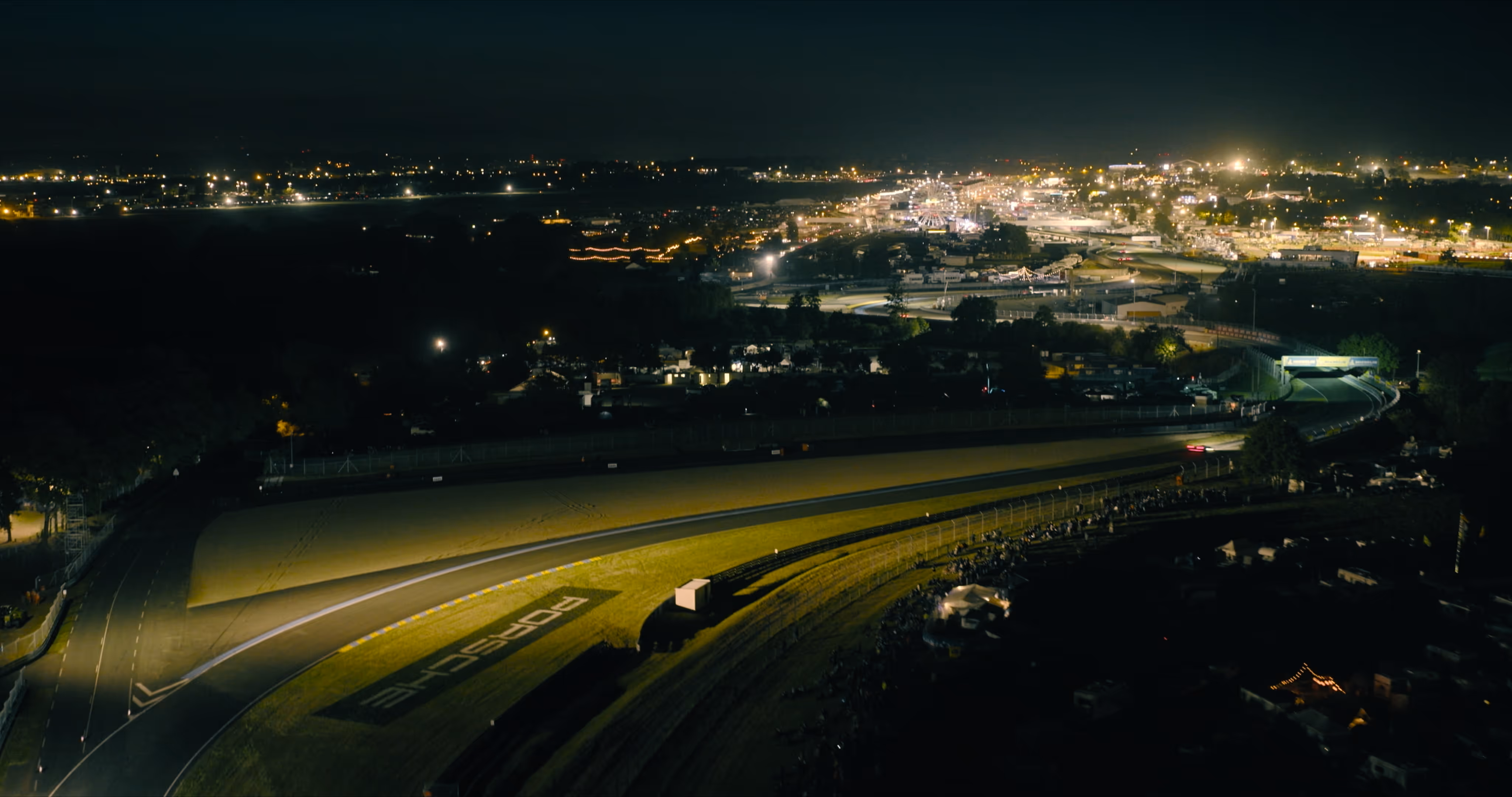 Aerial night view of a brightly lit race track with Porsche branding and surrounding illuminated cityscape.