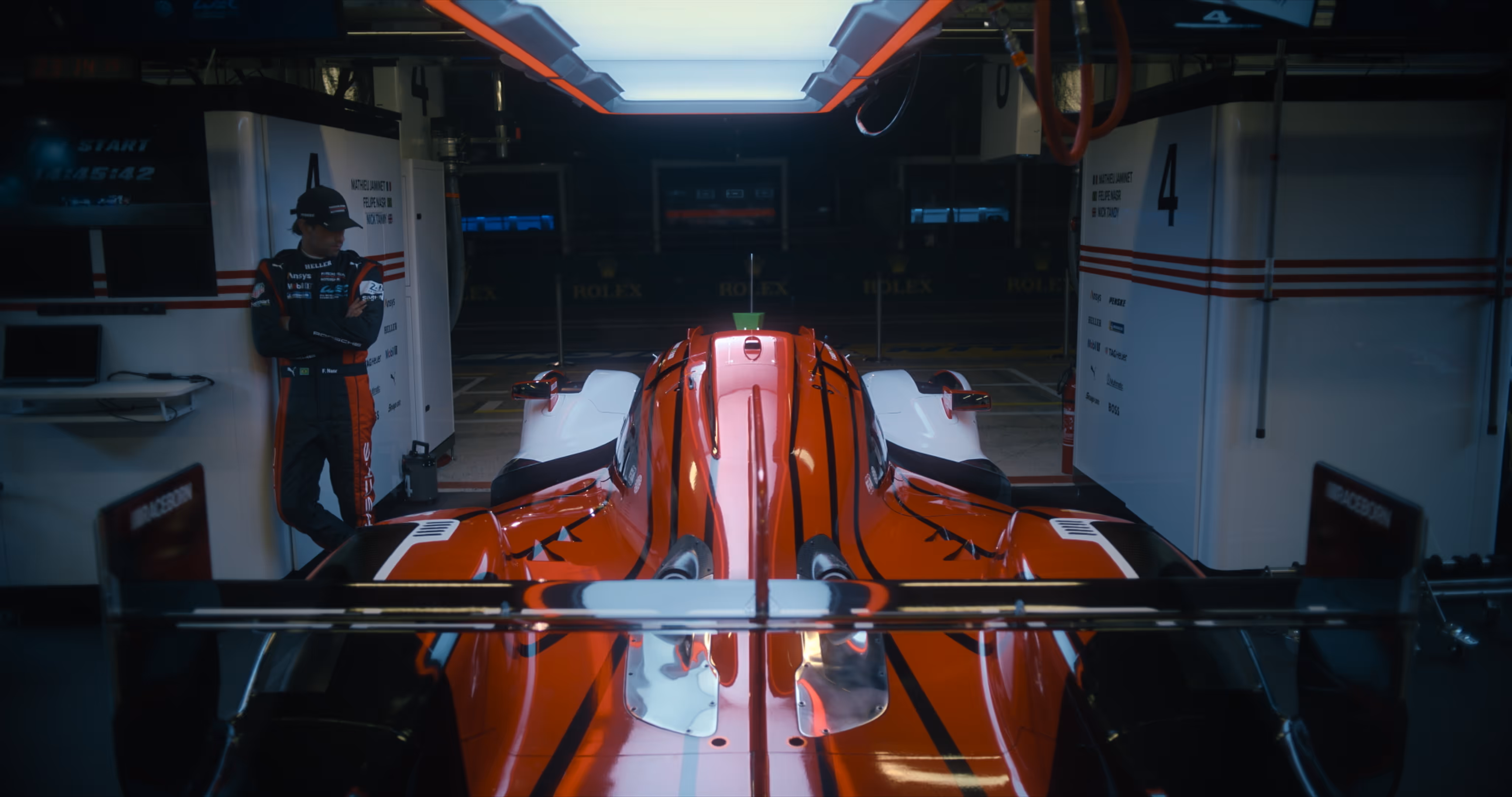 Rear view of a red and white racing car inside a dimly lit garage with a person in racing gear standing nearby.