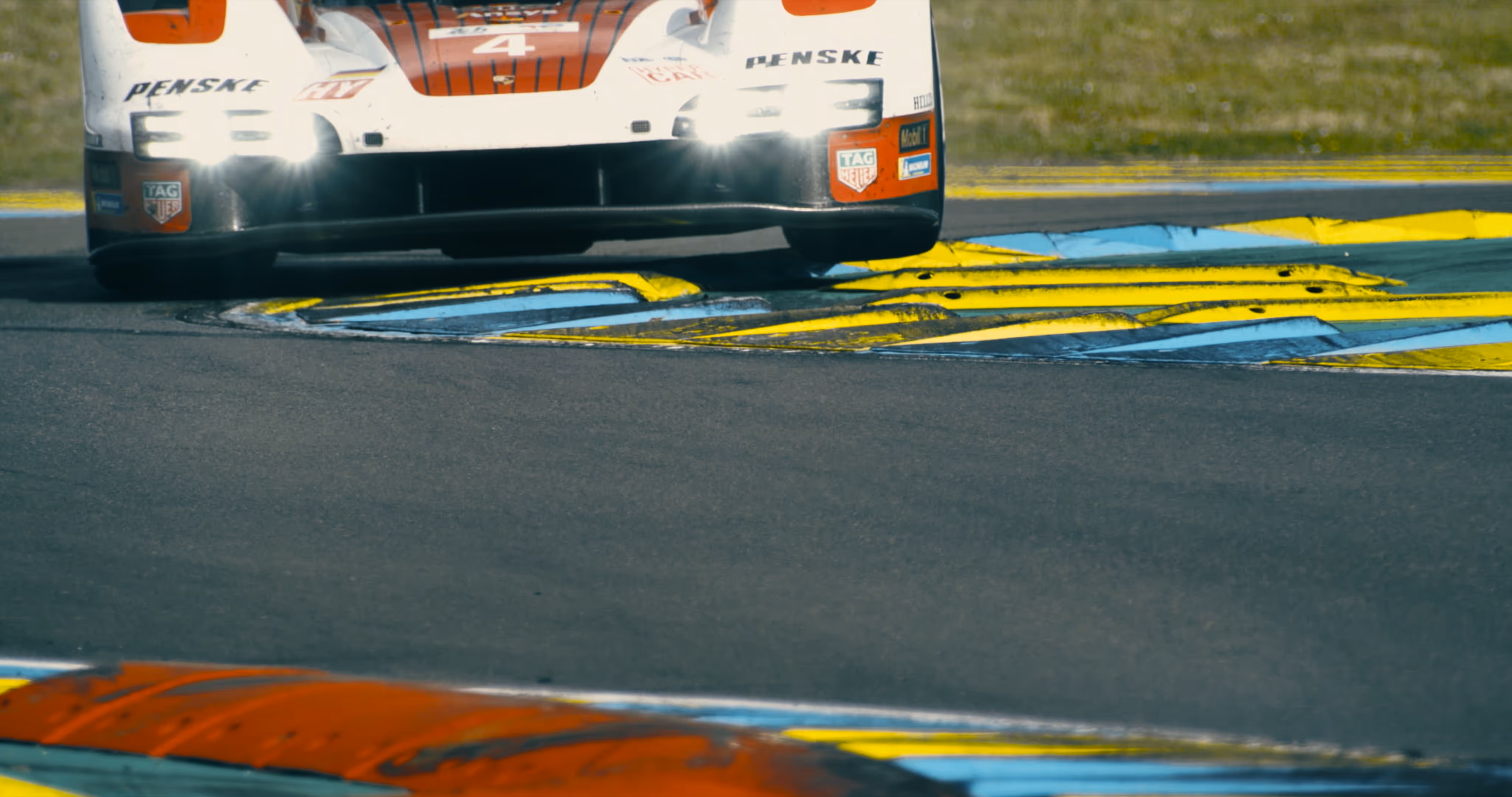 White and orange Penske race car with headlights on driving over blue and yellow curbs on a racetrack.